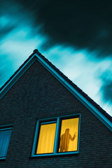 Intruder with handgun in eerie house with illuminated window under stormy sky at dusk.