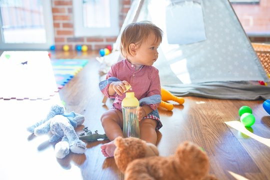 Adorable toddler holding feeding bottle around lots of toys at kindergarten