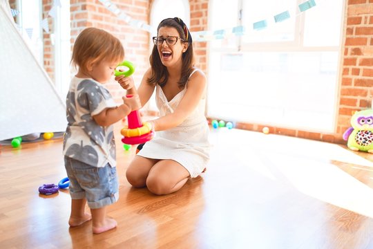 Beautiful teacher and toddler building pyramid using hoops around lots of toys at kindergarten