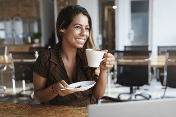 Woman enjoys tasty coffee in cafe during lunch. Attractive female entrepreneur working remote at co-working, hold cup and plate, smiling delighted as contemplate passersby and nature through window