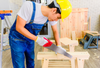 Asian young handyman and carpenter wearing yellow safety helmet and cutting wood by saw