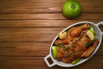 Baked whole chicken with apples and spices in the baking dish on wooden table with copy space. Horizontal top view.