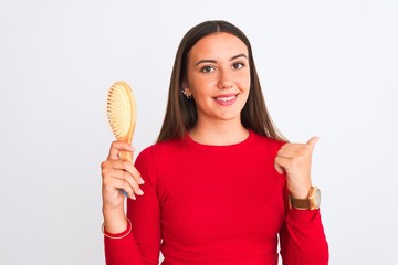 Young beautiful girl holding hair comb standing over isolated white background pointing and showing with thumb up to the side with happy face smiling