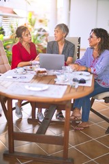 Meeting of middle age women having lunch and drinking coffee. Mature friends smiling happy using laptop at home on a sunny day