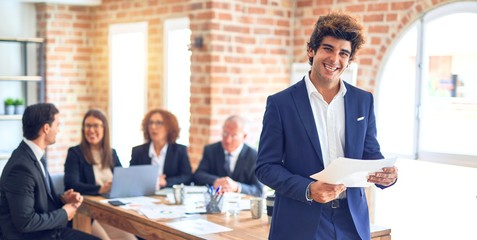 Group of business workers smiling happy and confident working together in a meeting. One of them, standing with smile on face reading documents at the office.