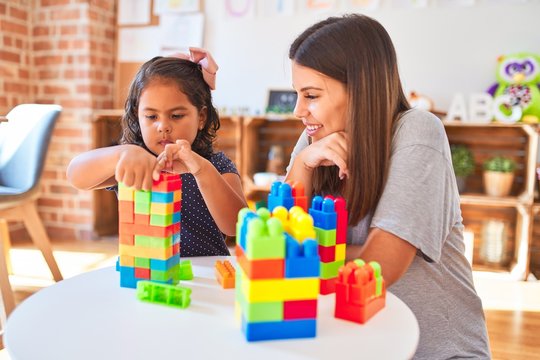 Beautiful teacher and toddler girl playing with construction blocks bulding tower at kindergarten