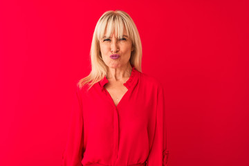 Middle age woman wearing elegant shirt standing over isolated red background puffing cheeks with funny face. Mouth inflated with air, crazy expression.