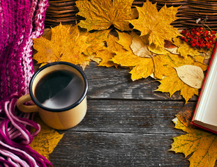 Hot coffee with steam in an enamel mug with a woolen scarf,  with an open book and leaves on wooden background. Autumn cozy background.