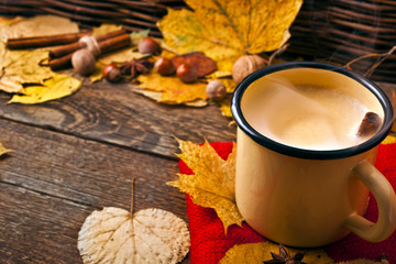 Coffee in an enamel mug with a woolen scarf, leaves, nuts, cinnamon sticks and star anise on wooden background. Autumn cozy background.