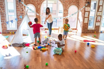 Young beautiful teacher and toddlers playing basketball around lots of toys at kindergarten