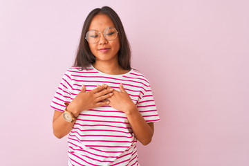 Young chinese woman wearing striped t-shirt and glasses over isolated pink background smiling with hands on chest with closed eyes and grateful gesture on face. Health concept.