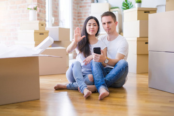 Fototapeta na wymiar Beautiful couple sitting on the floor using smartphone at new home around cardboard boxes with open hand doing stop sign with serious and confident expression, defense gesture