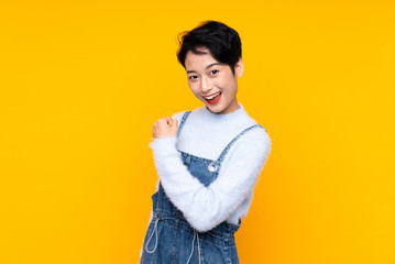 Young Asian girl in overalls over isolated yellow background celebrating a victory