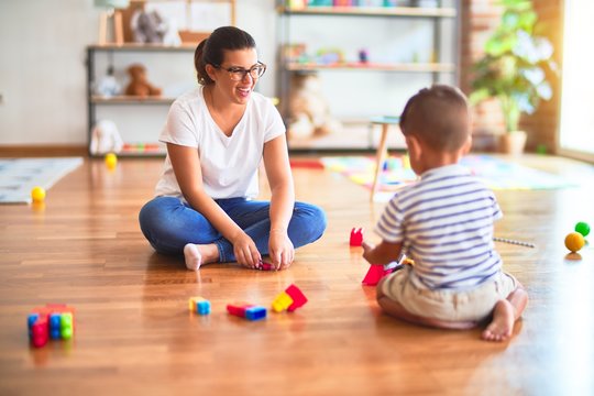 Beautiful teacher and toddler boy playing with tractor and cars at kindergarten