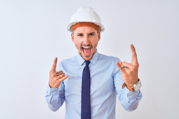 Young business man wearing contractor safety helmet over isolated background shouting with crazy expression doing rock symbol with hands up. Music star. Heavy concept.