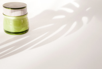 Cream in a green jar on a white background under the shadow of a tropical monstera plant. Eco cosmetics concept