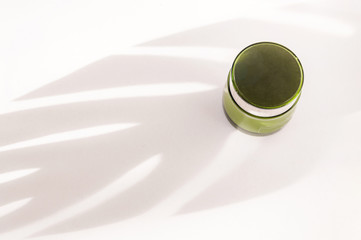 Cream in a green jar on a white background under the shadow of a tropical monstera plant. Eco cosmetics concept