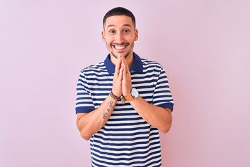 Young handsome man wearing nautical striped t-shirt over pink isolated background praying with hands together asking for forgiveness smiling confident.