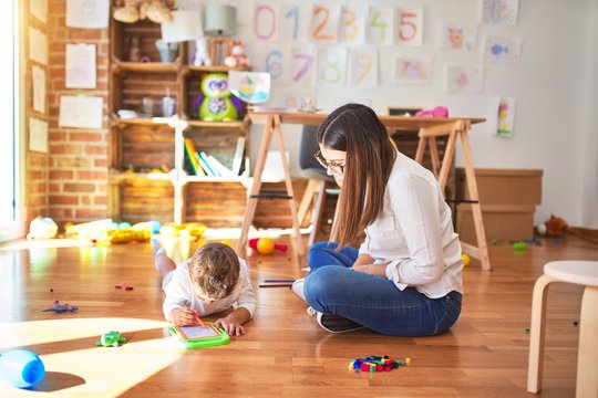 Beautiful teacher and toddler drawing using digital board around lots of toys at kindergarten