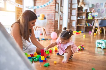 Young beautiful teacher and toddler sitting on the floor playing with building blocks toy at kindergarten