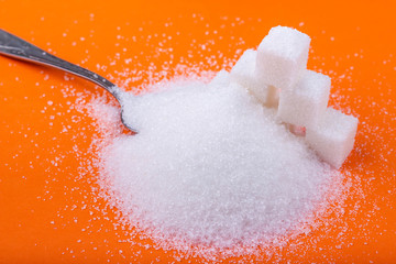 Spoon and cubes of white sugar and granulated sugar on an orange background