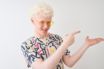 Young albino blond man wearing colorful t-shirt and sunglasses over isolated red background amazed and smiling to the camera while presenting with hand and pointing with finger.