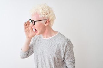 Young albino blond man wearing striped t-shirt and glasses over isolated white background shouting and screaming loud to side with hand on mouth. Communication concept.
