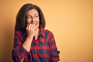 Middle age beautiful woman wearing casual shirt standing over isolated yellow background looking stressed and nervous with hands on mouth biting nails. Anxiety problem.