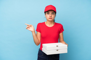 Young woman holding a pizza over isolated pink wall pointing to the laterals having doubts