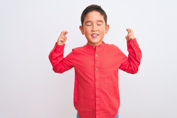 Beautiful kid boy wearing elegant red shirt standing over isolated white background gesturing finger crossed smiling with hope and eyes closed. Luck and superstitious concept.