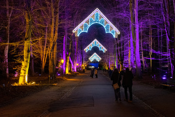 Park at night. Neon lighting decorations in shape of ornate arches in Christmas Garden Berlin. Orange and purple illuminated trees by sides of sidewalk in park. Holiday illuminations