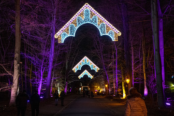 Neon lighting decorations in shape of ornate arches in Christmas Garden Berlin. Orange and purple illuminated trees by sides of sidewalk in park. Holiday illuminations