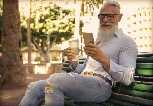 Trendy senior man using smartphone app and drinking coffee in the park. Mature fashion male having fun with new trends technology . Tech and joyful elderly lifestyle concept - Image