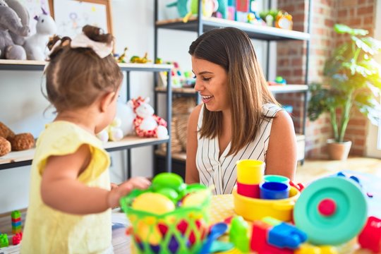 Young beautiful teacher and toddler playing with dishes, cutlery and cups toy on the table at kindergarten