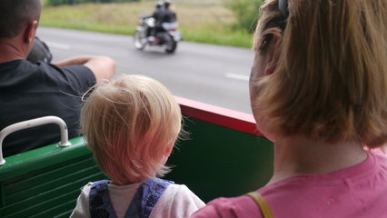 Kolejka wąskotorowa. Mama z dzieckiem jedzie pociągiem wśród zielonych łąk, w tle droga i przejeżdżający motocykl.  - obrazy, fototapety, plakaty