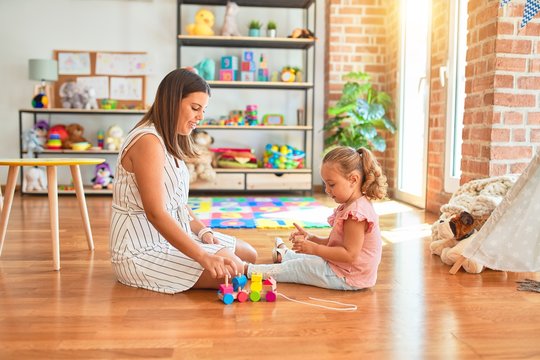 Beautiful teacher and blond student toddler girl playing with wooden train at kindergarten