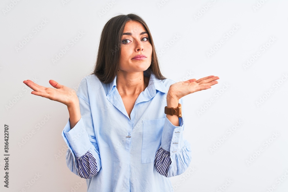 Wall mural Young beautiful woman wearing blue elegant shirt standing over isolated white background clueless and confused expression with arms and hands raised. Doubt concept.