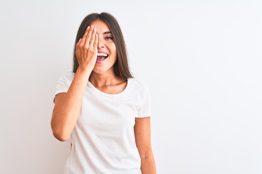 Young beautiful woman wearing casual t-shirt standing over isolated white background covering one eye with hand, confident smile on face and surprise emotion.