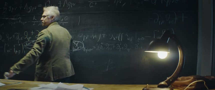 Gray haired Professor erases formula or equation from blackboard late at night. Senior scientist discovering formula, writes with chalk on blackboard. Anamorphic lens