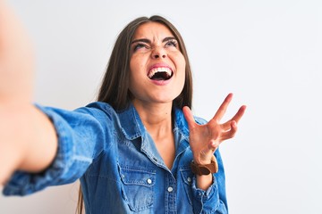 Obraz na płótnie Canvas Beautiful woman wearing denim shirt make selfie by camera over isolated white background crazy and mad shouting and yelling with aggressive expression and arms raised. Frustration concept.