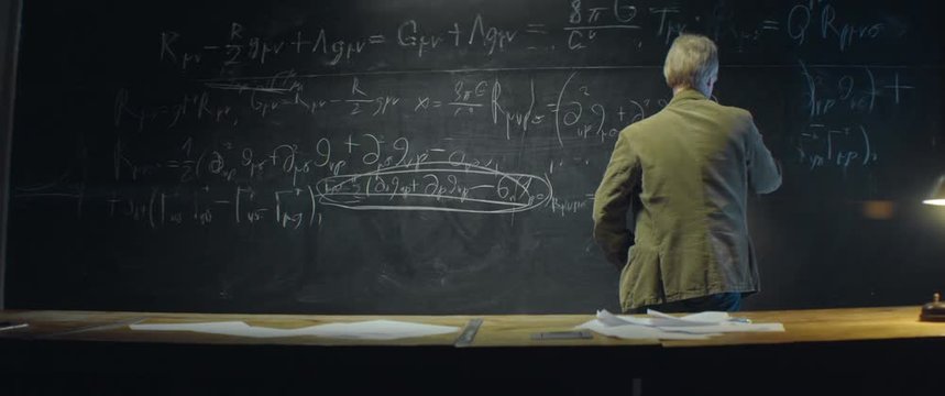 Aged scientist or Professor writing and erasing formula on blackboard late at night. University auditorium, table lamp shines. Anamorphic lens