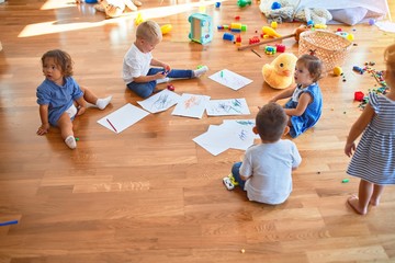 Adorable group of toddlers sitting on the floor drawing using paper and pencil around lots of toys...