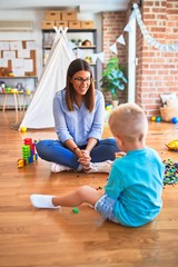 Young caucasian child playing at playschool with teacher. Mother and son at playroom bulding a tower with toy blocks