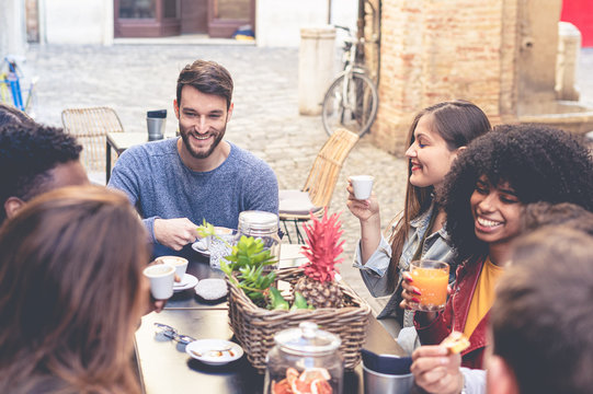 Group of young multiracial people having breakfast in an outdoor coffee shop. Students of different countries having fun together drinking coffees, juices and cappuccino sitting on a cafeteria table.