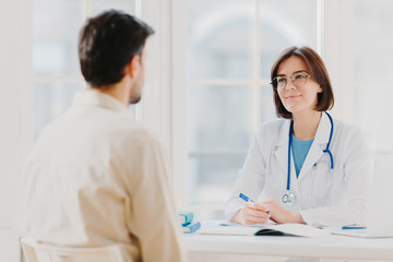 Doctor and patient discuss something, sit at table in clinic. Female cardiologist in eyewear gives...