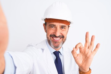 Middle age engineer man wearing helmet make selfie over isolated white background doing ok sign with fingers, excellent symbol