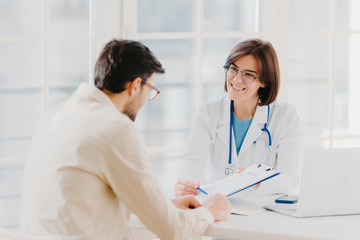 Woman doctor with stethoscope holds binder with patient personal medical card, consults patient who has medical problems, sit at hospital office, discuss medical checkup results, offers insurance
