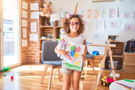 Beautiful toddler wearing glasses and unicorn diadem standing holding draw smiling at kindergarten