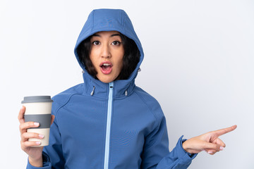 Mixed race woman wearing winter clothes with the hood on and holding a hot takeaway coffee over isolated white background surprised and pointing side