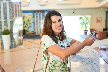 Young beautiful woman smiling cheerful using smartphone standing on hotel hall on holidays